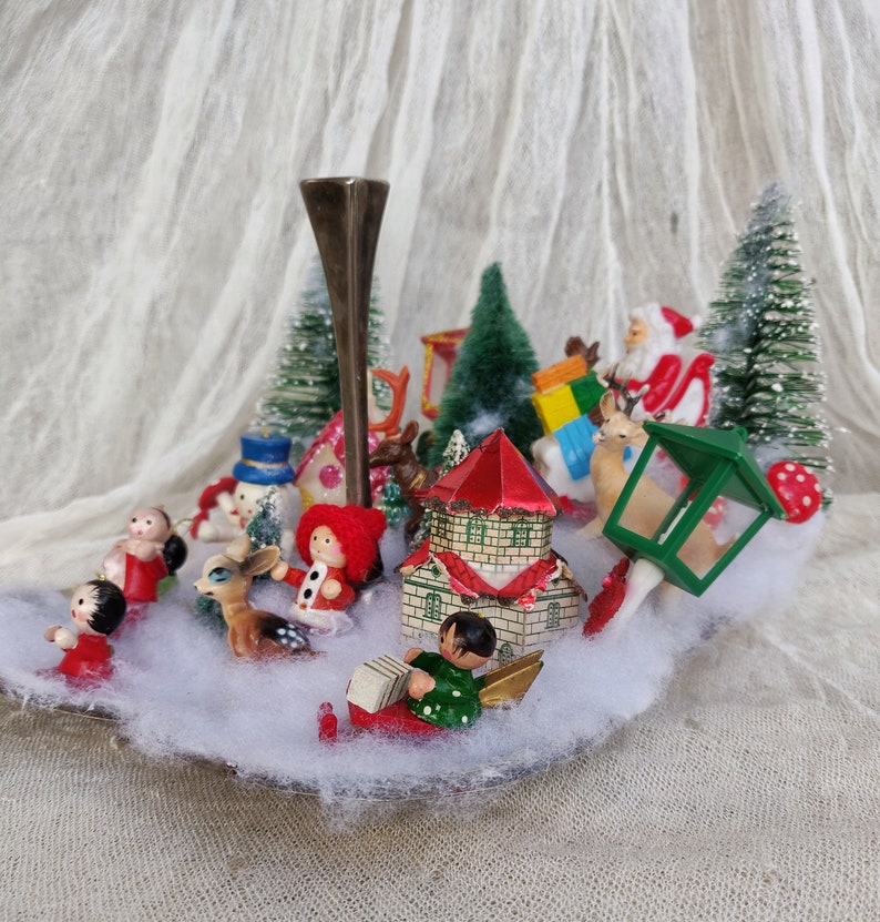 May include: A festive Christmas display featuring miniature figurines of Santa, a snowman, and children, surrounding a central candle holder. The scene includes small houses, trees, and a lantern, set on a bed of artificial snow.