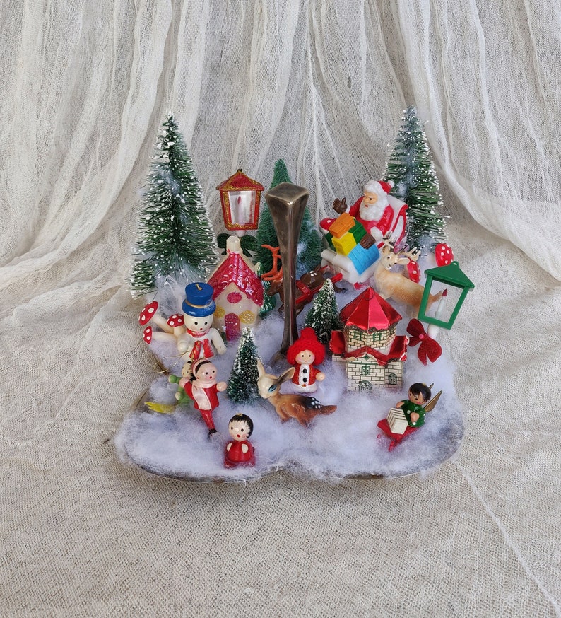 May include: A vintage Christmas diorama with miniature figures of Santa, reindeer, children, and snowmen. The scene includes small houses, trees, and street lamps, set against faux snow. The colour scheme is dominated by red, green, and white, evoking a festive atmosphere.