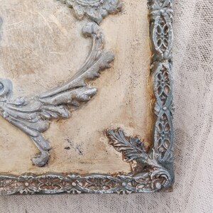 May include: Close-up of a decorative wall panel with ornate, raised relief details. The panel features a distressed, off-white background with gray-blue accents on the decorative frame and floral motifs. The corner of the panel is visible.