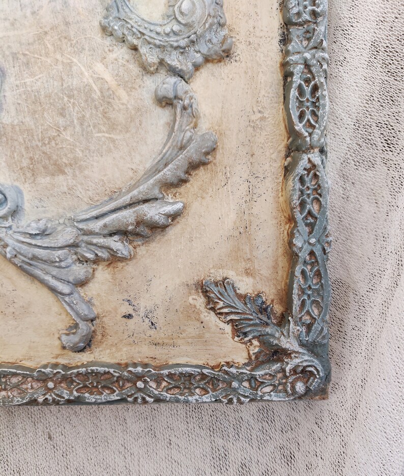 May include: Close-up of a decorative wall panel with ornate, raised relief details. The panel features a distressed, off-white background with gray-blue accents on the decorative frame and floral motifs. The corner of the panel is visible.
