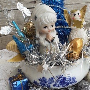 May include: A vintage Christmas centerpiece featuring a porcelain figurine, a blue tinsel tree, and an angel figurine. The arrangement is set in a white teacup with blue floral patterns, adorned with silver tinsel, gold and silver ornaments, and a small blue gift box.