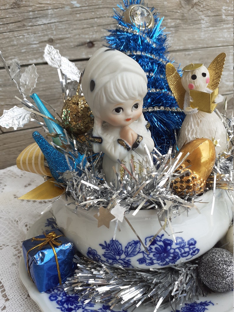 May include: A vintage Christmas centerpiece featuring a porcelain figurine, a blue tinsel tree, and an angel figurine. The arrangement is set in a white teacup with blue floral patterns, adorned with silver tinsel, gold and silver ornaments, and a small blue gift box.