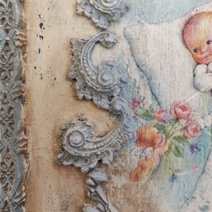 May include: A vintage-style decorative plaque with a painted image of a baby holding flowers. The plaque features ornate, light gray scrollwork and a light blue border. The background has a distressed, aged appearance, with the word "Christening" visible.