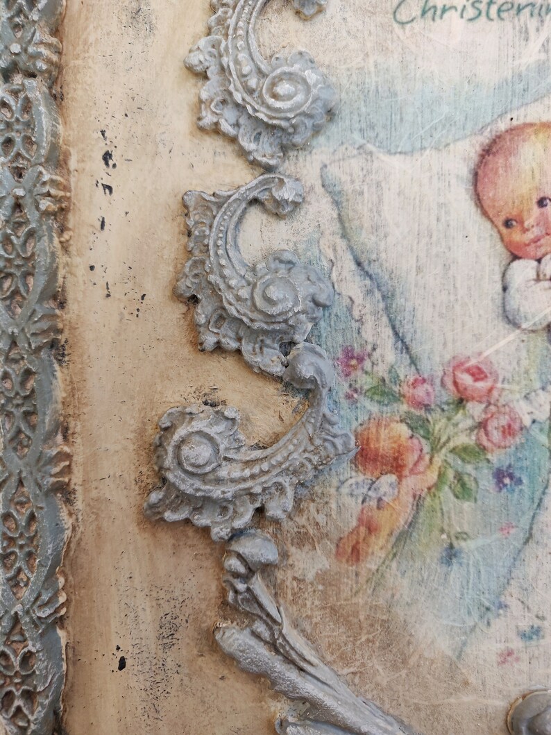 May include: A vintage-style decorative plaque with a painted image of a baby holding flowers. The plaque features ornate, light gray scrollwork and a light blue border. The background has a distressed, aged appearance, with the word "Christening" visible.