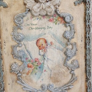 May include: A vintage-style plaque with a baby illustration, framed by ornate, light blue scrollwork and a praying angel. The text "On Your Christening Day" is visible. The background is a cream color with a distressed finish, giving it a rustic appearance.