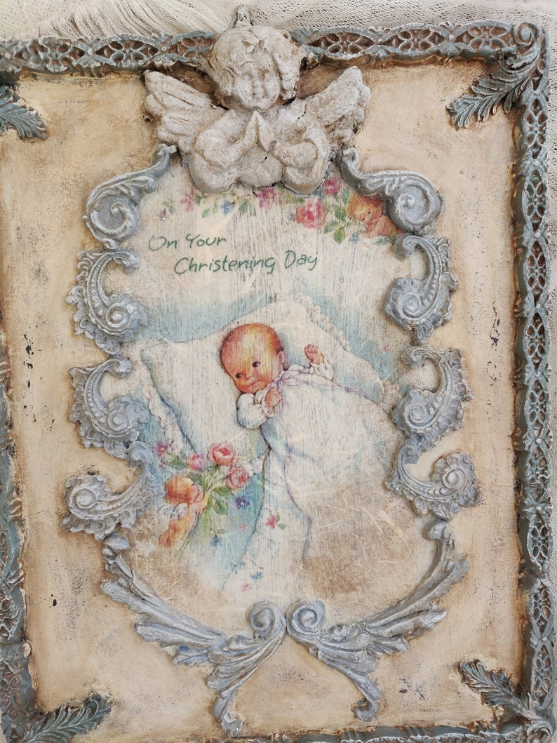 May include: A vintage-style plaque with a baby illustration, framed by ornate, light blue scrollwork and a praying angel. The text "On Your Christening Day" is visible. The background is a cream color with a distressed finish, giving it a rustic appearance.