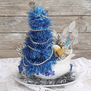 May include: A festive Christmas decoration featuring a blue tinsel tree adorned with a rhinestone garland, set in a white gravy boat with blue floral patterns. Silver tinsel and decorative elements complete the holiday arrangement.