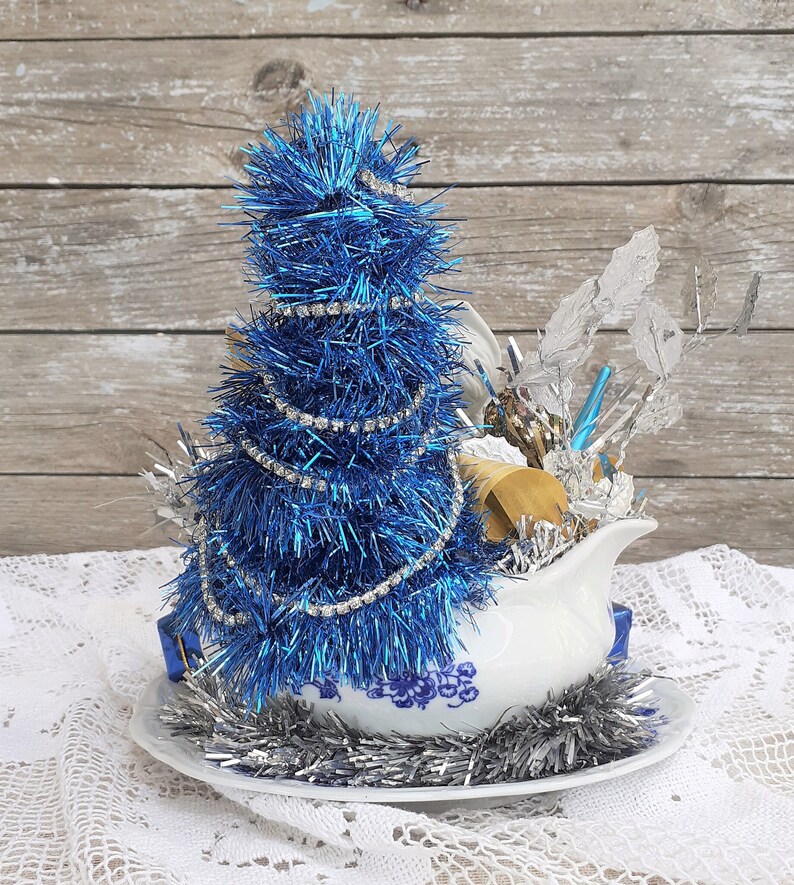 May include: A festive Christmas decoration featuring a blue tinsel tree adorned with a rhinestone garland, set in a white gravy boat with blue floral patterns. Silver tinsel and decorative elements complete the holiday arrangement.