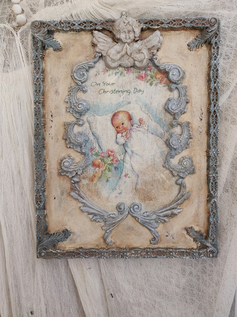 May include: A vintage-style framed artwork with a baby illustration, angel, and floral accents. The frame is distressed with a blue-gray finish. The artwork includes the text "On Your Christening Day".