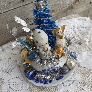 May include: A vintage Christmas ornament arrangement featuring a blue tinsel tree, porcelain figurines, and silver tinsel. The arrangement is set in a blue and white porcelain bowl with a floral design, and includes small gift boxes.