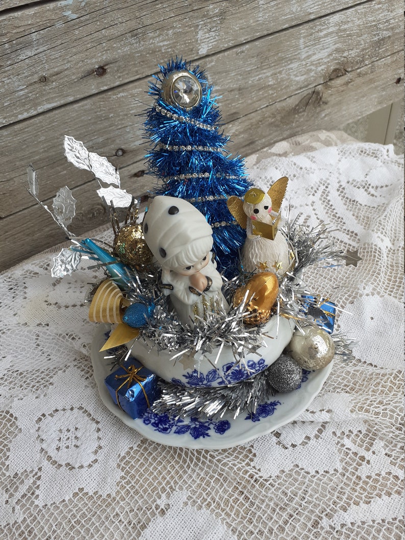 May include: A vintage Christmas ornament arrangement featuring a blue tinsel tree, porcelain figurines, and silver tinsel. The arrangement is set in a blue and white porcelain bowl with a floral design, and includes small gift boxes.
