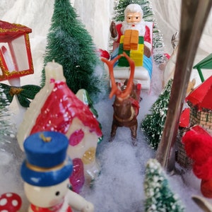 May include: A miniature Christmas village scene with a snowman, Santa Claus in a sleigh pulled by a reindeer, and small houses. The scene includes miniature green trees and a snowy landscape, creating a festive holiday display.