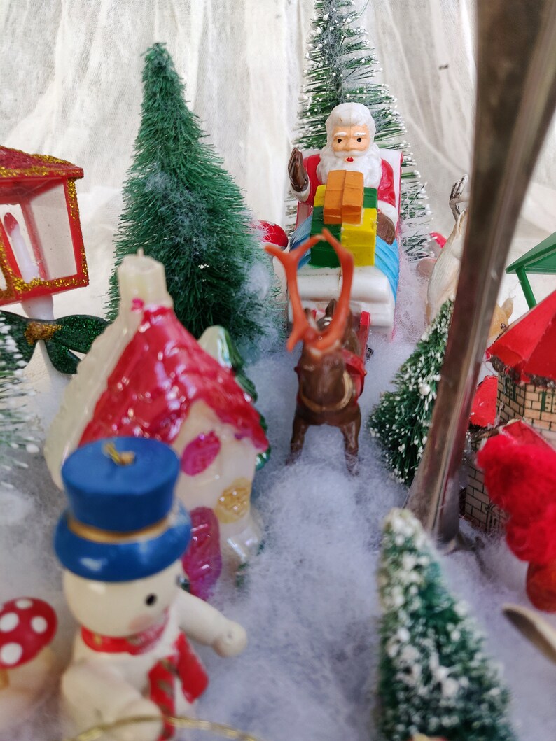 May include: A miniature Christmas village scene with a snowman, Santa Claus in a sleigh pulled by a reindeer, and small houses. The scene includes miniature green trees and a snowy landscape, creating a festive holiday display.