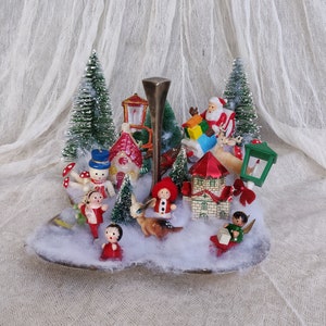 May include: A vintage Christmas display with miniature figurines of Santa, reindeer, snowmen, and children. The scene includes bottle brush trees, houses, and a decorative metal stand, set against a backdrop of faux snow.