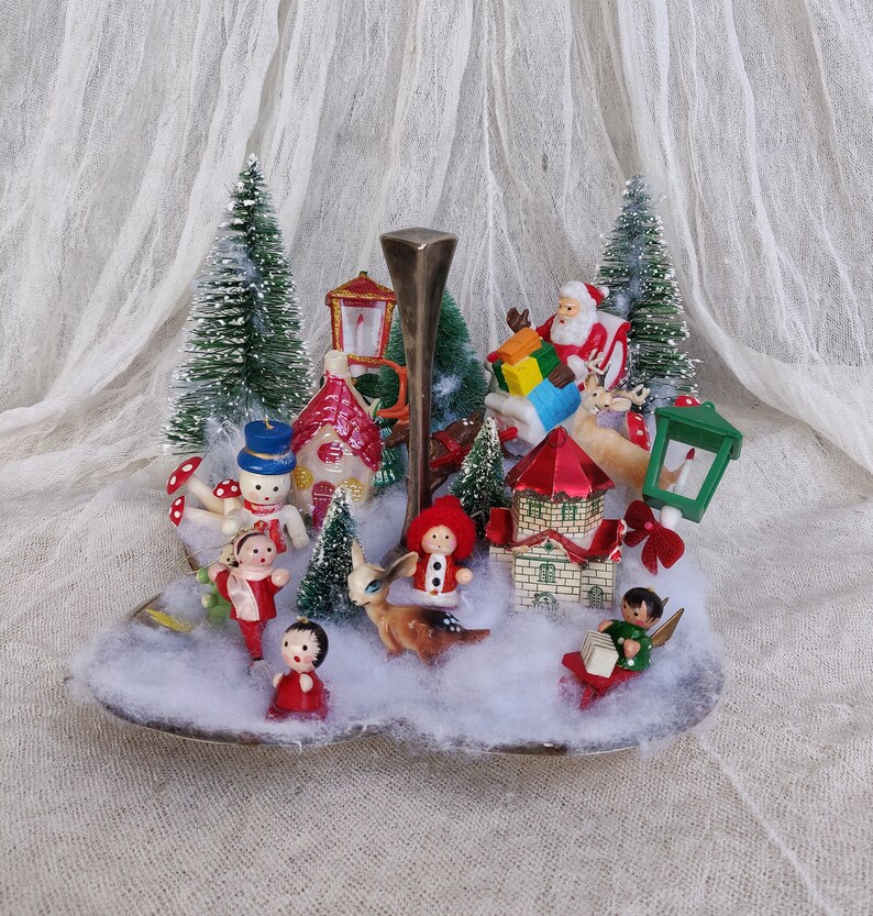 May include: A vintage Christmas display with miniature figurines of Santa, reindeer, snowmen, and children. The scene includes bottle brush trees, houses, and a decorative metal stand, set against a backdrop of faux snow.