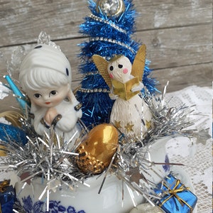 May include: A festive Christmas arrangement featuring a blue tinsel tree, porcelain figurines of angels and a girl, and various ornaments. The arrangement is set in a white porcelain dish with blue floral patterns, accented with silver tinsel.