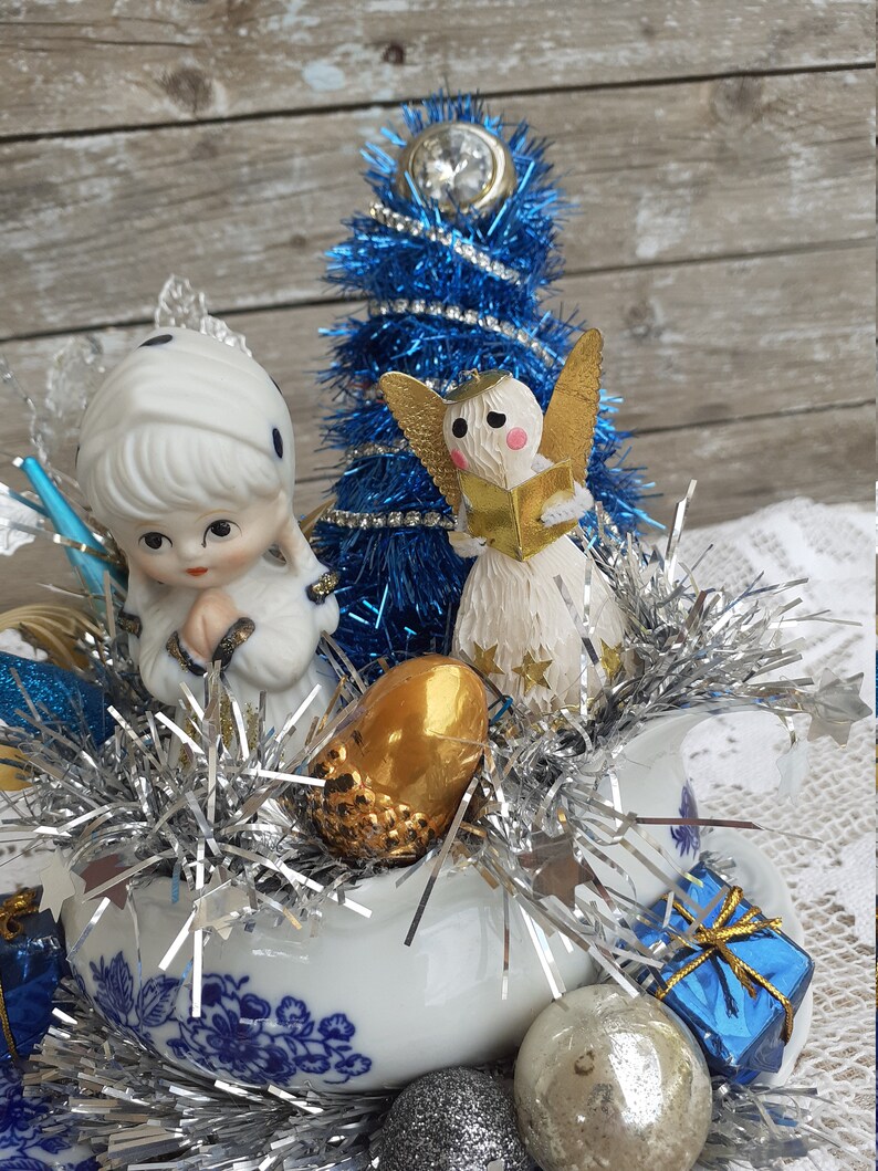 May include: A festive Christmas arrangement featuring a blue tinsel tree, porcelain figurines of angels and a girl, and various ornaments. The arrangement is set in a white porcelain dish with blue floral patterns, accented with silver tinsel.