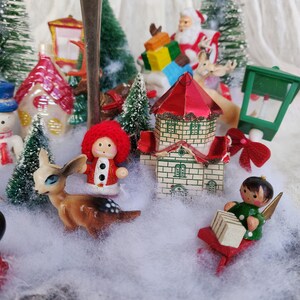 May include: A Christmas-themed miniature display featuring Santa Claus, a reindeer, a snowman, and a small house. Miniature Christmas trees and faux snow surround the figurines, all set on a silver base.