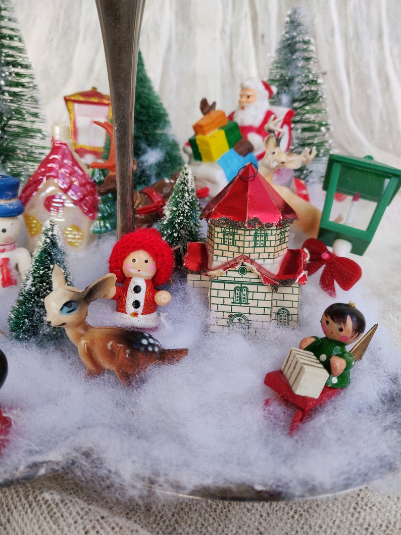 May include: A Christmas-themed miniature display featuring Santa Claus, a reindeer, a snowman, and a small house. Miniature Christmas trees and faux snow surround the figurines, all set on a silver base.