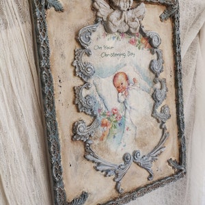 May include: A vintage-style rectangular wall plaque with a distressed finish. The plaque features a central image of a baby with the text "On Your Christening Day". The frame is adorned with ornate, gray-blue scrollwork and a small angel figurine at the top.