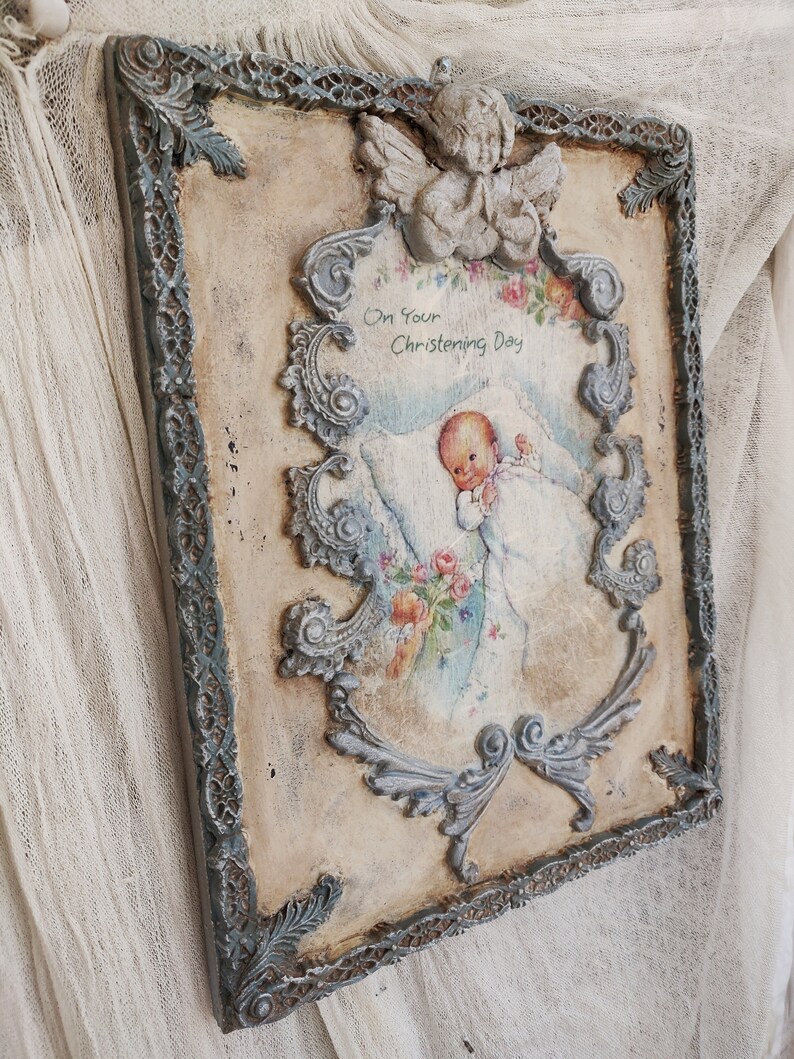 May include: A vintage-style rectangular wall plaque with a distressed finish. The plaque features a central image of a baby with the text "On Your Christening Day". The frame is adorned with ornate, gray-blue scrollwork and a small angel figurine at the top.
