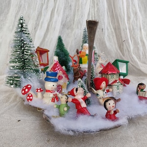 May include: A miniature Christmas village scene with bottle brush trees, houses, and wooden figurines of Santa, reindeer, and children. The scene is set on a silver spoon with faux snow and red and white mushroom decorations.