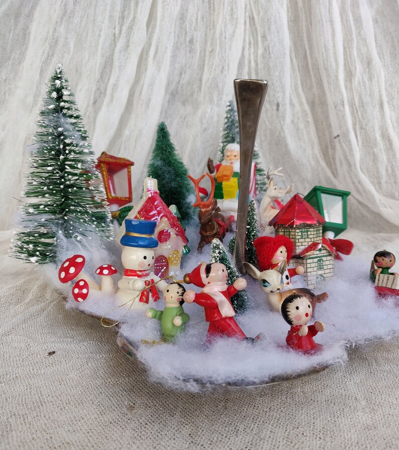 May include: A miniature Christmas village scene with bottle brush trees, houses, and wooden figurines of Santa, reindeer, and children. The scene is set on a silver spoon with faux snow and red and white mushroom decorations.