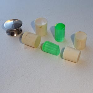 May include: A collection of small cylinders and a silver knob. The cylinders are in shades of cream and green. The silver knob has a rounded top and a decorative base. The objects are arranged on a white surface.