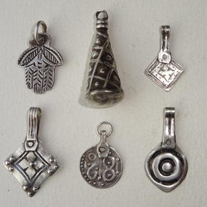 Könnte beinhalten: Eine Sammlung von sechs silberfarbenen Anhängern mit verschiedenen Designs. Die Anhänger umfassen eine Hamsa-Hand, eine Kegelform, zwei Diamantenformen, einen runden Anhänger mit einem geätzten Design und einen Anhänger mit einem kreisförmigen Design.