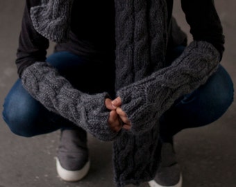 May include: A gray knitted winter set, including a hat with a pom-pom, a scarf, and fingerless gloves. The set features a cable knit design. The person is wearing a black long-sleeve shirt and blue jeans.