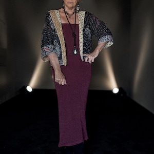 May include: A burgundy sleeveless midi dress paired with a black and cream patterned open-front jacket. The jacket features three-quarter length sleeves and a contrasting cream-coloured trim along the front edges. The outfit is accessorised with a long beaded necklace.