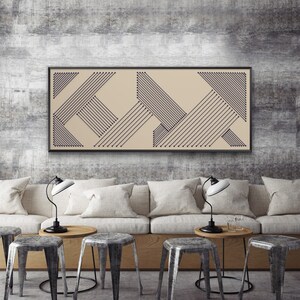 Puede incluir: Una obra de arte abstracta enmarcada con un fondo beige y líneas geométricas azul oscuro. La obra es rectangular y cuelga sobre un sofá beige con cojines blancos. La habitación también cuenta con taburetes de metal y mesas de madera.