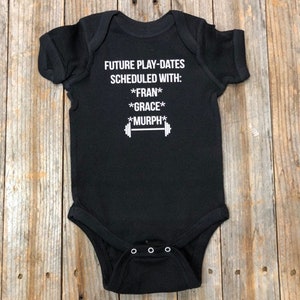 Op de afbeelding: Zwarte romper met witte tekst die "Future Play-Dates Scheduled With: *Fran* *Grace* *Murph*" luidt en een halterstang afbeelding.