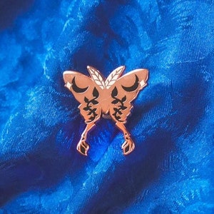 Könnte beinhalten: Roségoldene Schmetterlingsnadel mit schwarzen Halbmonden und floralen Details auf einem blauen Stoffhintergrund.