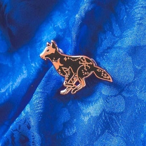 Puede incluir: Un pin de esmalte dorado con un lobo negro con detalles celestiales, incluyendo estrellas y una luna creciente. El pin está sobre un fondo de tela azul.