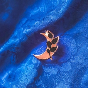 Puede incluir: Un pin de media luna de oro rosa con un diseño de hoja negra. El pin está sobre un fondo de tela azul.