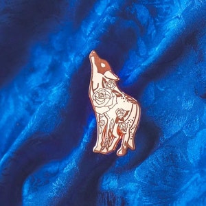 Puede incluir: Un pin de esmalte de oro rosa con un lobo blanco aullando a la luna. El lobo está rodeado de rosas blancas. El pin está sobre un fondo de tela azul.