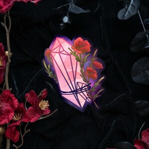 May include: A die-cut sticker featuring a pink, faceted crystal point wrapped in dark blue cord, adorned with vibrant red poppy flowers and green stems, set against a dark purple and black gradient background with sharp, defined edges.