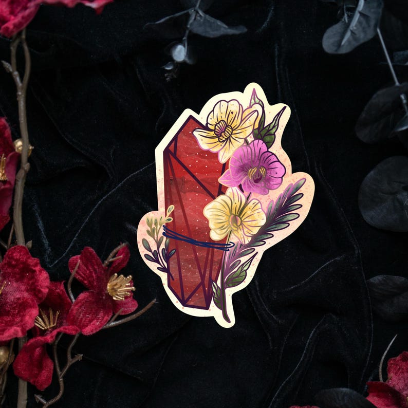 Puede incluir: Una pegatina troquelada que muestra una punta de cristal facetada de color rojo intenso envuelta en un cordel oscuro, adornada con flores amarillas y rosas, hojas verdes y peque&ntilde;as ramitas, con un borde color crema sobre fondo oscuro.