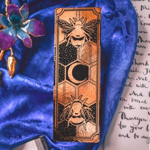 Puede incluir: Una caja rectangular de color cobre con un diseño negro que presenta dos abejas, una luna creciente y patrones de panal. La caja está decorada con un fondo estrellado y enmarcada por un borde decorativo.