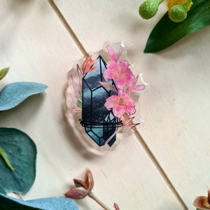 Puede incluir: Un amuleto de resina con un cristal azul y blanco con un contorno negro, rodeado de flores rosas y hojas verdes.