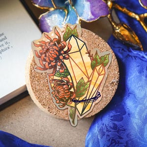 May include: A die-cut sticker featuring two yellow, geometric crystals intertwined with orange chrysanthemum flowers and green leaves. The illustration has a clear outline and a subtle holographic sheen, showcasing a botanical and mineral design.