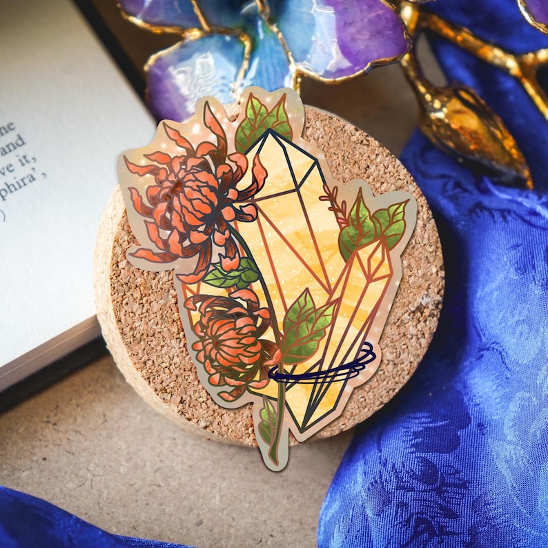 May include: A die-cut sticker featuring two yellow, geometric crystals intertwined with orange chrysanthemum flowers and green leaves. The illustration has a clear outline and a subtle holographic sheen, showcasing a botanical and mineral design.