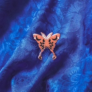 Puede incluir: Un broche de mariposa color cobre con detalles intrincados, incluyendo formas de media luna y patrones estilizados. El broche se encuentra sobre una tela azul oscuro texturizada, creando un contraste llamativo.