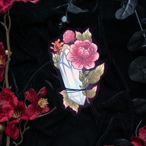 May include: A sticker featuring a vertical, light-colored crystal point wrapped in a thin dark cord, adorned with large pink peony blossoms and green leaves. The illustration has a dark outline and is set against a deep, dark background.