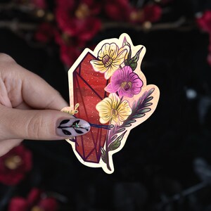 Puede incluir: Una pegatina troquelada que presenta una forma de cristal geom&eacute;trico de color rojo intenso adornada con un racimo de flores amarillas y moradas, hojas verdes y un acento de cinta azul oscuro, sobre un borde de fondo color crema.