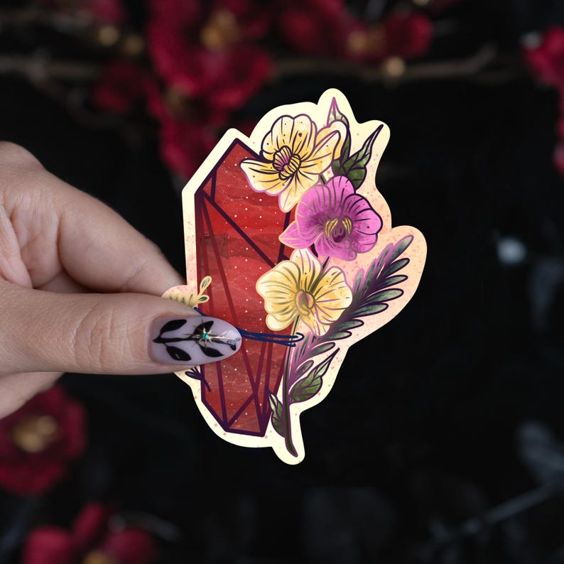 Puede incluir: Una pegatina troquelada que presenta una forma de cristal geom&eacute;trico de color rojo intenso adornada con un racimo de flores amarillas y moradas, hojas verdes y un acento de cinta azul oscuro, sobre un borde de fondo color crema.
