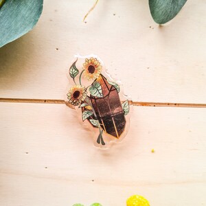 Puede incluir: Un amuleto de resina transparente con un cristal marrón y girasoles. El cristal es facetado y tiene un efecto de purpurina dorada. Los girasoles son amarillos con centros marrones.