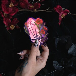 May include: A die-cut sticker featuring a pink and purple geometric crystal illustration intertwined with vibrant red poppy flowers and green stems. The design has a dark purple outline and a glossy finish, showcasing a blend of floral and mineral elements.