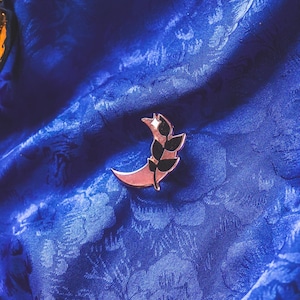 Puede incluir: Un broche en forma de media luna con un diseño de estrella y hojas. El broche es de color oro rosa con detalles en negro, sobre un fondo de tela azul floral.